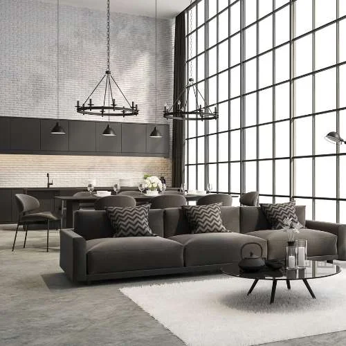 A combined living and dining space with matching gray sofas, pillows, and a striking light fixture, all under an open high ceiling and framed by glass walls in metal frames.