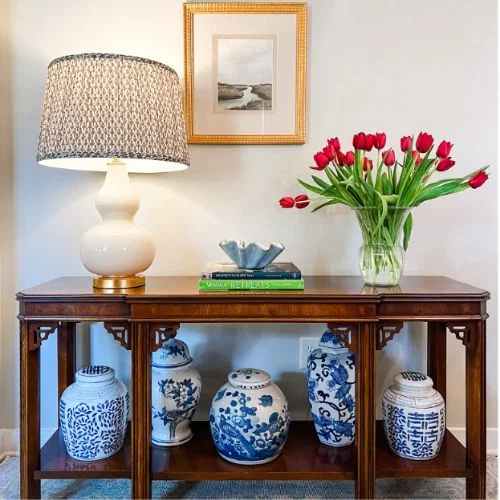 A classic, timeless wooden console entryway table decor with carved fretwork, adorned by blue-and-white porcelain vases, a lamp, and fresh tulips on top.