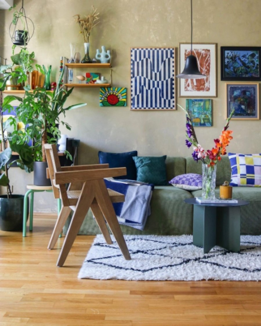 Eclectic bohemian living room with olive green sofa, wood chairs, colorful art, and plants.