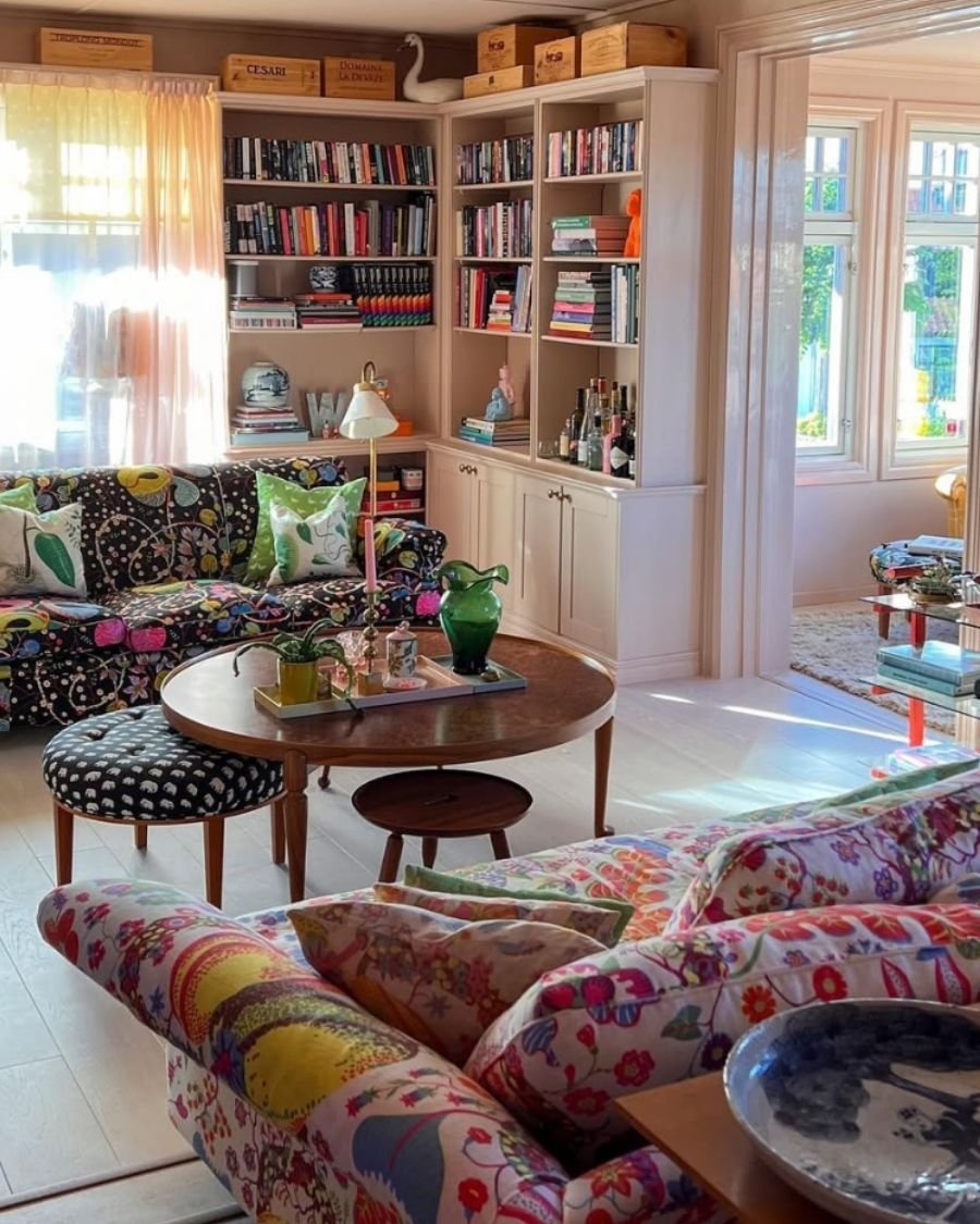 Eclectic library corner with black patterned sofa and red floral sofa, green pillows, built-in shelves, and pink candles.