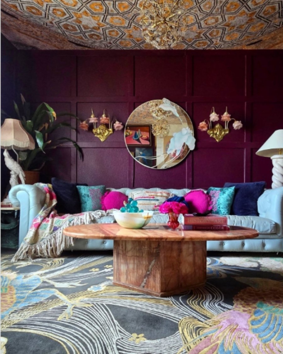 Eclectic living room with burgundy walls, blue tufted sofa, bold pillows, and patterned rug.