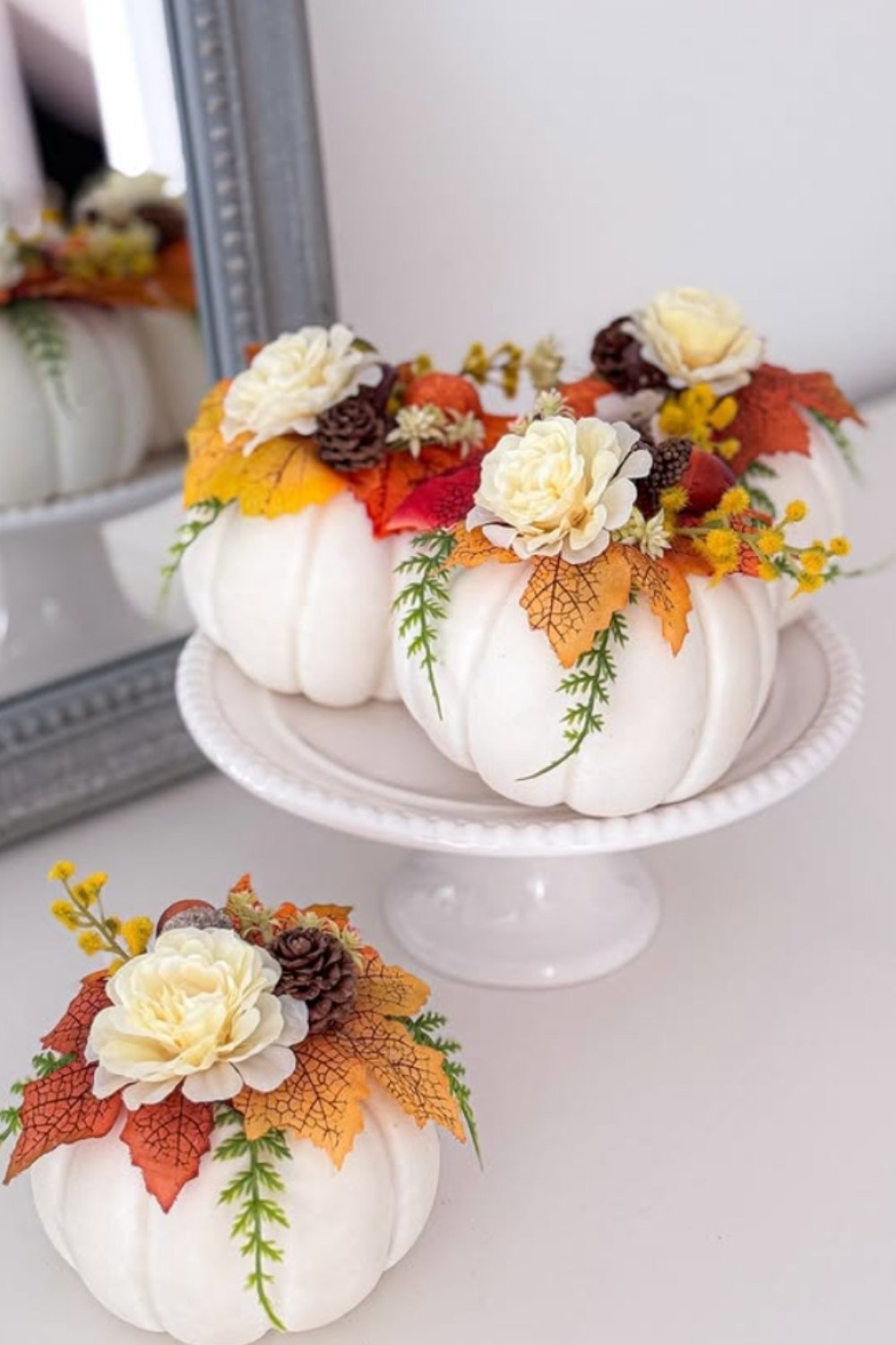 White pumpkin floral arrangements displayed on a cake stand with roses, mums, and autumn leaves.