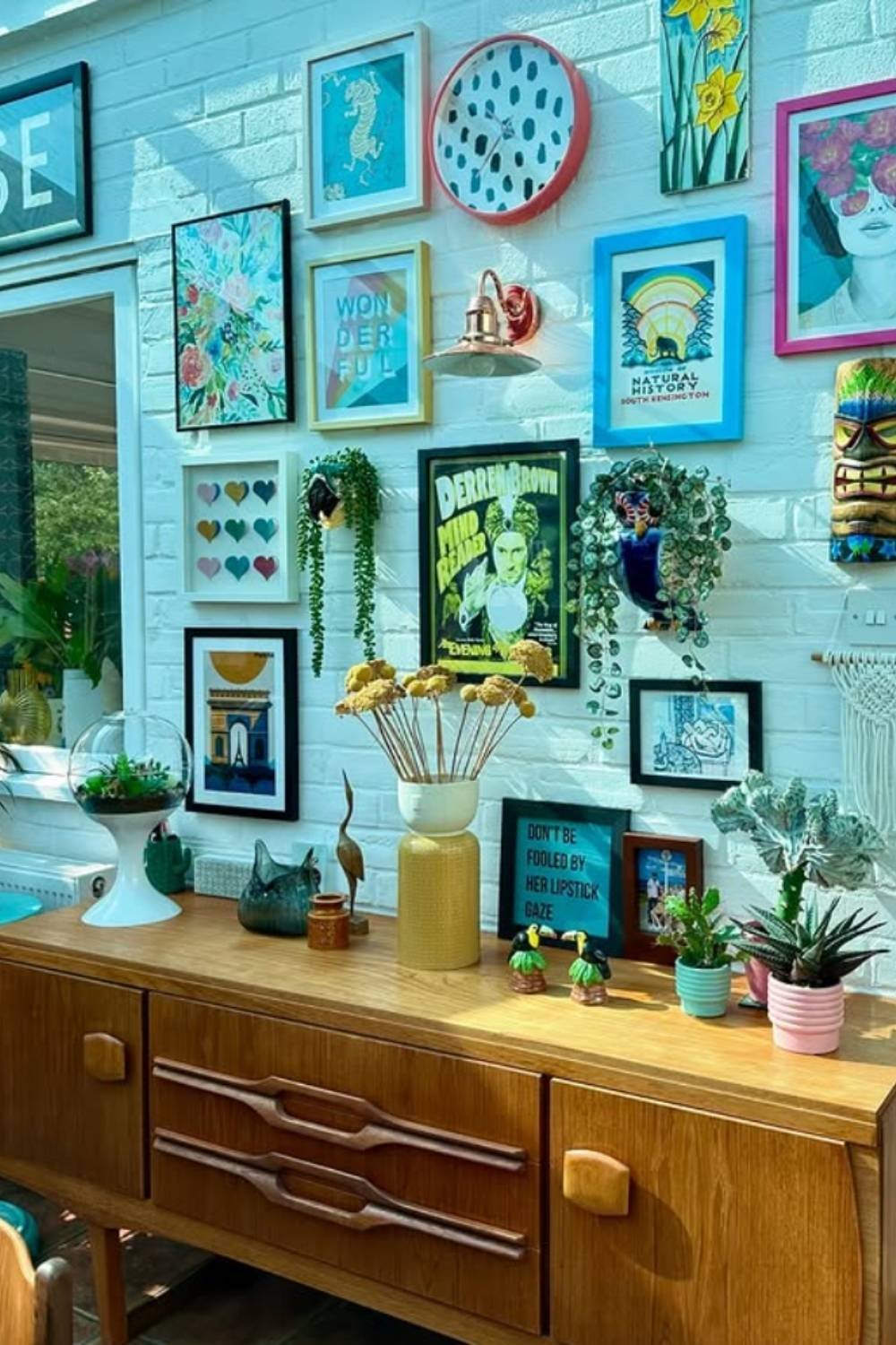 Colorful gallery wall with turquoise frames above a wooden sideboard and plants.