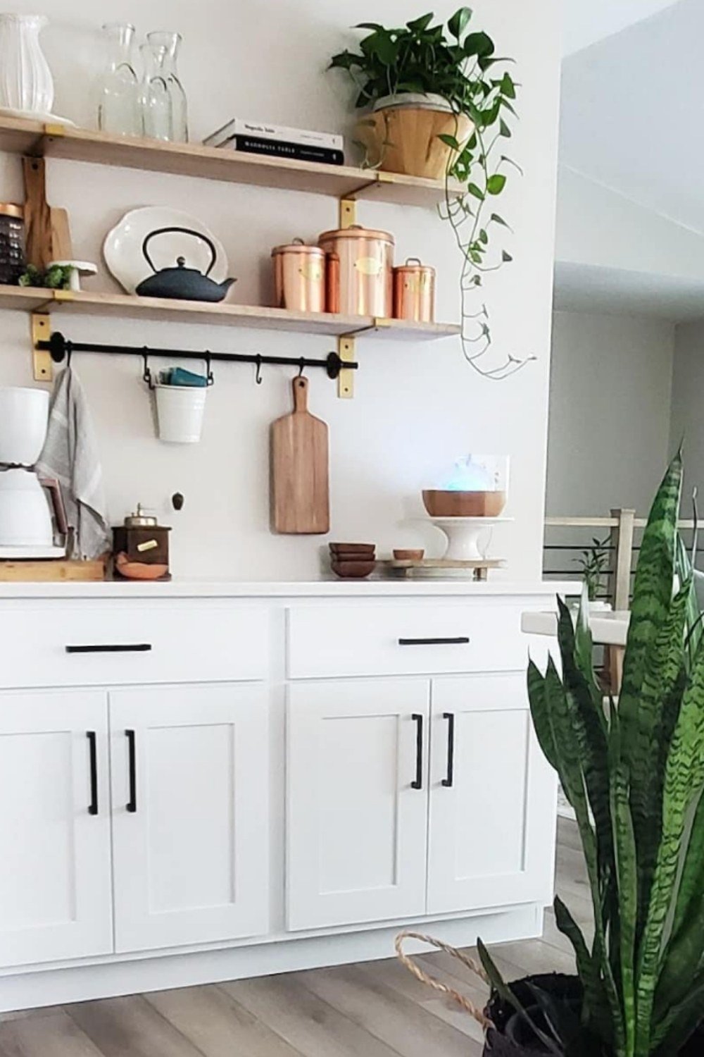 White cabinet coffee bar with copper jars, cutting boards, and hanging mugs.
