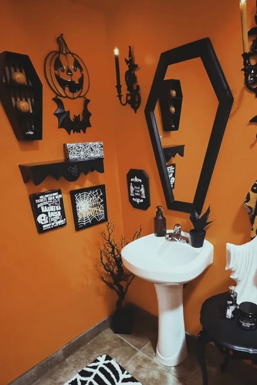 Pumpkin and bat-decorated Halloween bathroom with orange walls, framed spooky art, and a coffin-shaped mirror.